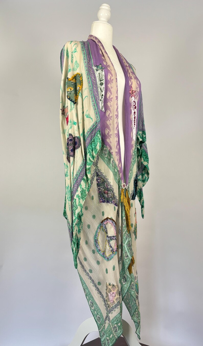 May include: A long, flowing kimono in cream, green, and purple. The kimono features various patches with designs like hearts, peace signs, and the word "LOVE". It has a decorative tassel and is displayed on a mannequin.