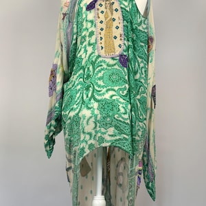 May include: A long, flowing kimono-style garment in cream and green paisley print. The back features an embroidered patch with a figure in a gold dress. Purple butterfly appliques and trim add detail to this unique piece.