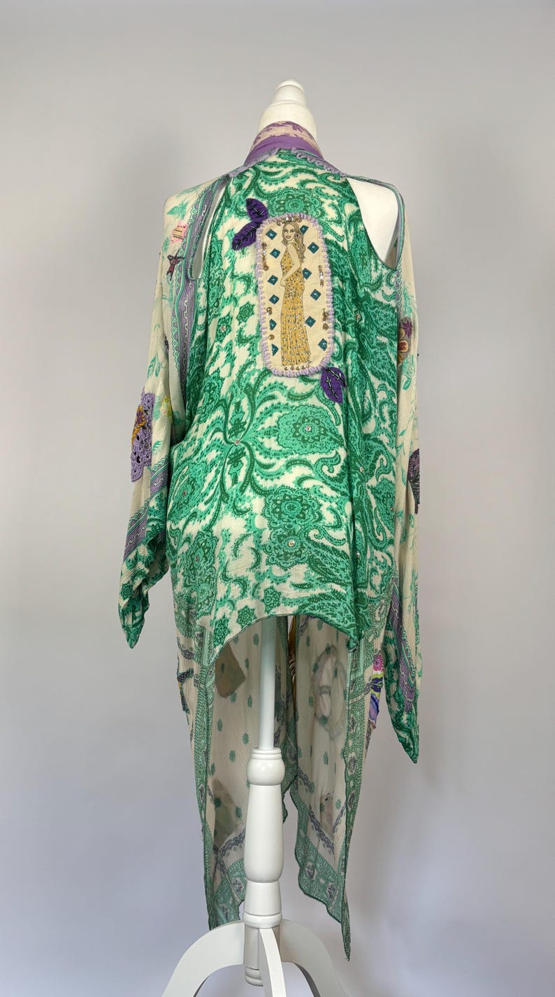 May include: A long, flowing kimono-style garment in cream and green paisley print. The back features an embroidered patch with a figure in a gold dress. Purple butterfly appliques and trim add detail to this unique piece.