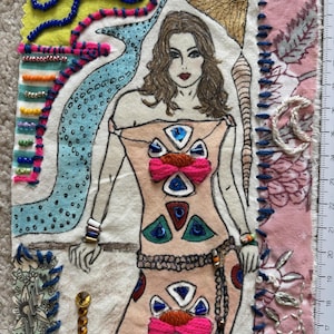May include: Textile art piece featuring a woman in a peach-colored dress with pink bows and geometric shapes. The artwork includes beaded embellishments, embroidery, and a blue and pink decorative border. A ruler is visible on the right side.