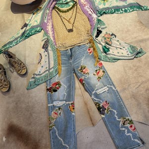 May include: An ensemble of women's clothing including light wash denim jeans with floral embroidery and distressed details, a gold-colored lace top, a patterned kimono, and layered necklaces. A wide-brimmed hat and sandals complete the look.