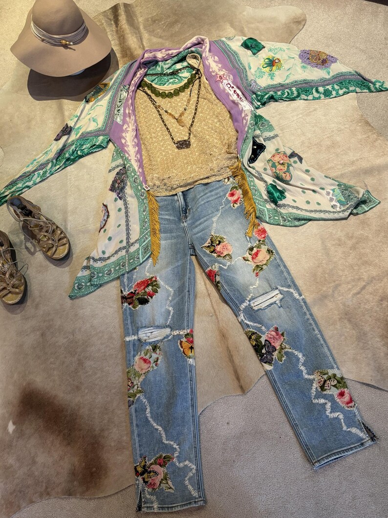 May include: An ensemble of women's clothing including light wash denim jeans with floral embroidery and distressed details, a gold-colored lace top, a patterned kimono, and layered necklaces. A wide-brimmed hat and sandals complete the look.