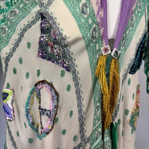 May include: A cream-colored, patterned kimono-style garment with green and purple accents. The garment features decorative patches and a purple collar with gold fringe and floral embellishments. The fabric has a sheer quality.