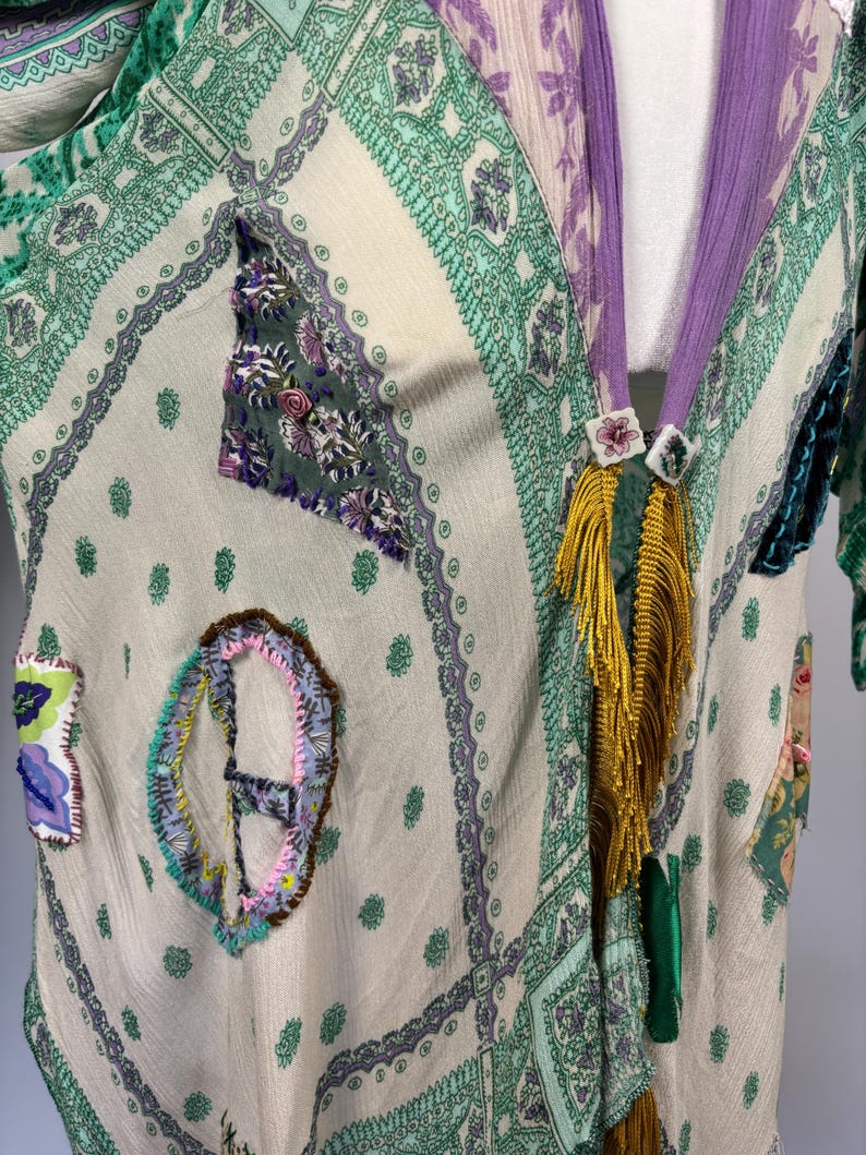 May include: A cream-colored, patterned kimono-style garment with green and purple accents. The garment features decorative patches and a purple collar with gold fringe and floral embellishments. The fabric has a sheer quality.