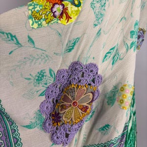 May include: A cream-colored garment with floral and leaf patterns in teal and green. It features decorative patches with floral designs in various colors, including yellow, purple, and orange. The edges are trimmed with a purple and green patterned border.