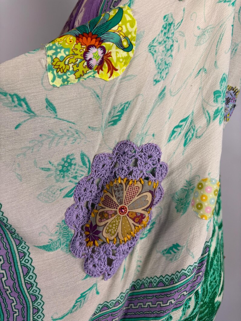 May include: A cream-colored garment with floral and leaf patterns in teal and green. It features decorative patches with floral designs in various colors, including yellow, purple, and orange. The edges are trimmed with a purple and green patterned border.