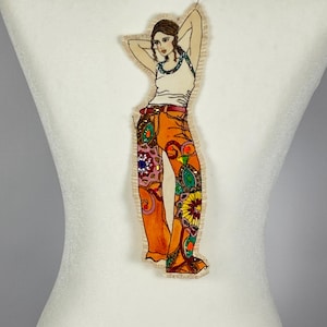 May include: A fabric applique of a woman in a white tank top and orange patterned pants. The woman has her arms behind her head. The pants have a variety of colorful floral and geometric designs. The applique is attached to a white surface.