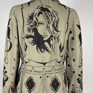 May include: A khaki-colored jacket featuring a large black graphic illustration of a person's face on the back. The jacket is decorated with black painted floral patterns, stars, and diamond-shaped patches accented with small black studs and brass rivets.