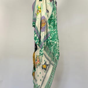 May include: A cream-colored, patterned kimono-style garment with green paisley and floral designs. The garment features patches with star and floral motifs, and is trimmed with gold tassels. The kimono is displayed on a white mannequin.