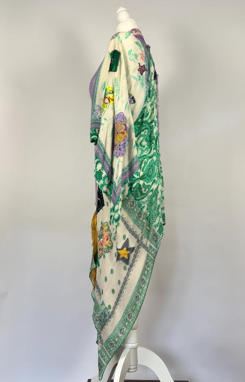 May include: A cream-colored, patterned kimono-style garment with green paisley and floral designs. The garment features patches with star and floral motifs, and is trimmed with gold tassels. The kimono is displayed on a white mannequin.
