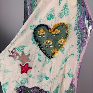 May include: Cream-colored fabric with a floral and leaf pattern, accented with a heart-shaped patch. The patch is green with yellow and silver details. Additional star and floral patches in pink, red, and purple add to the design. The edges are trimmed with a purple and green border.