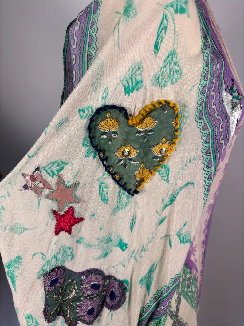 May include: Cream-colored fabric with a floral and leaf pattern, accented with a heart-shaped patch. The patch is green with yellow and silver details. Additional star and floral patches in pink, red, and purple add to the design. The edges are trimmed with a purple and green border.