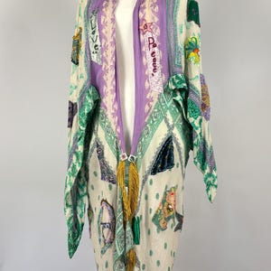 May include: A long, open-front garment with a cream background, adorned with patches of various colors and patterns. The garment features a purple and cream striped collar and a gold tassel. The sleeves and edges are trimmed with green patterned fabric.
