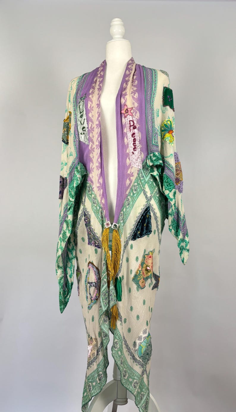 May include: A long, open-front garment with a cream background, adorned with patches of various colors and patterns. The garment features a purple and cream striped collar and a gold tassel. The sleeves and edges are trimmed with green patterned fabric.