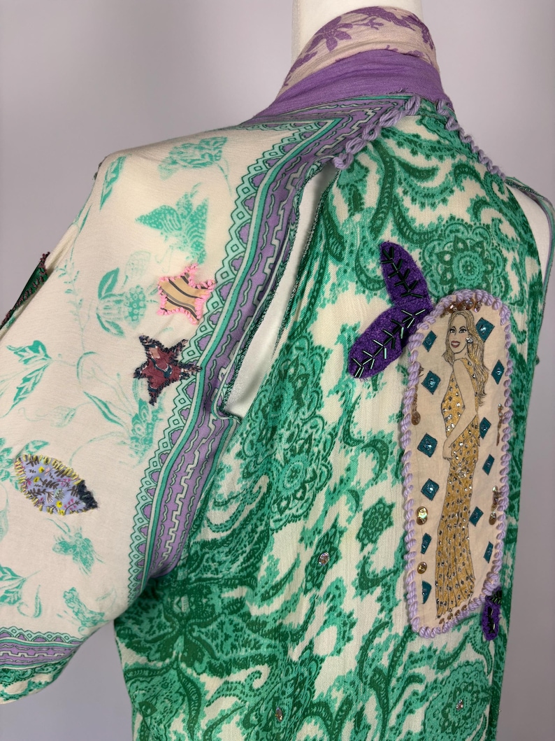 May include: A patterned, long-sleeved garment with a green and white paisley design. The garment features a purple collar and trim, with decorative patches and embellishments. A patch with a woman in a gold dress is on the back.