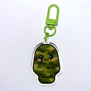 Peut inclure: Un porte-clés en acrylique transparent avec un personnage de dessin animé souriant au motif camouflage. Le porte-clés a un fermoir vert et une finition pailletée, avec des joues roses et un contour noir.