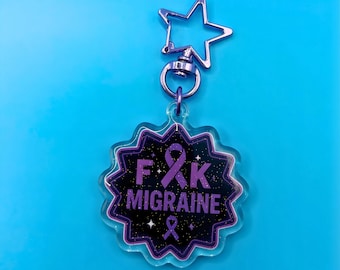 May include: A clear acrylic keychain with a star-shaped clasp. The keychain features a black and purple design with the text "F*K MIGRAINE" and a purple ribbon symbol. The keychain is on a blue background.