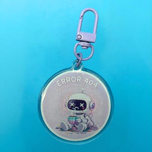 May include: A round keychain with a lilac clasp. The keychain features a cartoon robot with the text "ERROR 404. Motivation Not Found." The robot is sitting and holding a cup, with a sad expression.