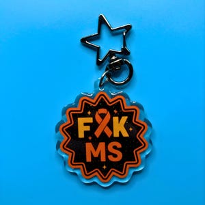 May include: A clear acrylic keychain with a star-shaped clasp. The keychain has a black and orange design with the letters "F&K MS" and an orange ribbon. The background is a solid blue.