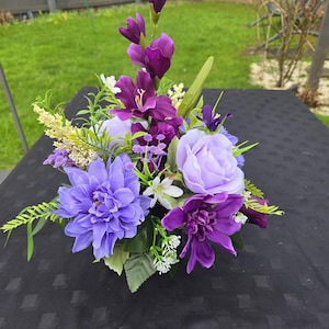 May include: A colourful floral display with purple and lilac blooms, such as dahlias, roses, and gladioli. The arrangement includes small white flowers and green leaves, presented on a black surface.