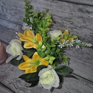 May include: A bouquet of yellow and white artificial flowers with green leaves. The flowers are arranged in a cluster and are sitting on a wooden surface.