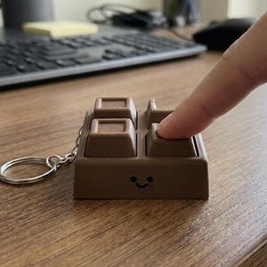May include: A brown, square-shaped fidget keychain designed to resemble a chocolate bar. The object features four raised, clickable square buttons on its top surface and a small, simple black smiley face printed on the front side.