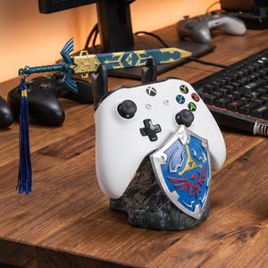 May include: A white video game controller resting on a dark, textured rock-shaped stand. The stand features a blue and silver shield emblem on the front and a miniature blue and gold sword with a long blue tassel attached to the top of the base.