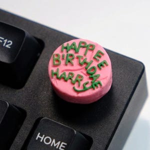 May include: A pink, circular, decorative item with green text that reads "HAPPY BIRTHDAY HARRIE" sits on a black keyboard. The item resembles a small cake or candy, adding a festive touch to the keyboard.