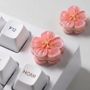 May include: A white keyboard with two pink flower-shaped keycaps. The keycaps have a layered design with yellow centres. The keyboard keys include F12, SPACE, and HOM.