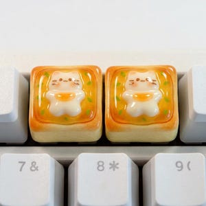 Può includere: Due tasti per tastiera personalizzati con un design a forma di gatto su toast. I tasti sono quadrati, con una combinazione di colori giallo e arancione, e sono circondati da tasti bianchi della tastiera. Il gatto è bianco con dettagli arancioni.