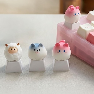 Op de afbeelding: Een verzameling aangepaste toetsenbordtoetsen met dierontwerpen. De toetsen omvatten een wit schaap, een blauw-witte kat en twee roze en witte kattenkappen. De toetsen liggen op een wit oppervlak en een roze toetsenbord.