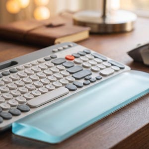May include: A wireless computer keyboard featuring a light gray frame, white circular keys with dark gray accents, and a single orange circular key. The keyboard includes a translucent, light blue gel wrist rest attached to the front edge.