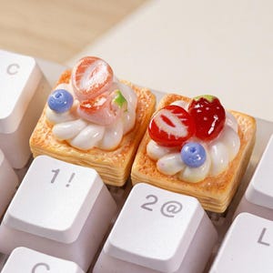 May include: Two decorative keycaps shaped like miniature pastries, each topped with whipped cream, strawberries, and blueberries. The keycaps are on a white keyboard, with the "1!" and "2@" keys visible.