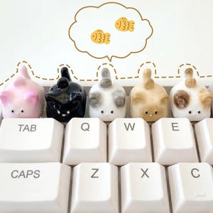 Puede incluir: Un conjunto de cinco teclas con forma de gato en varios colores, incluyendo rosa, negro, blanco, beige y carey, colocadas en un teclado blanco. Una burbuja de pensamiento con dos ilustraciones de peces amarillos flota encima.