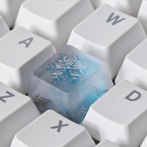 Könnte beinhalten: Nahaufnahme einer Computertastatur mit weißen Tasten. Eine Taste ist ein durchscheinender blauer Würfel mit einem Schneeflockenmuster. Die Schneeflocke ist hellblau. Die anderen Tasten sind weiß mit grauen Buchstaben.