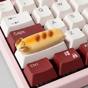 May include: A mechanical keyboard with white and burgundy keycaps. A novelty keycap shaped like a loaf of bread rests on the keyboard. The keyboard has a pink base.