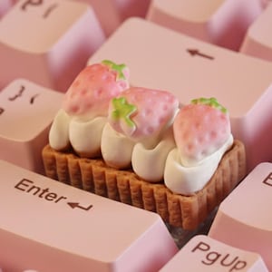 May include: A pink mechanical keyboard with a custom keycap shaped like a strawberry tart. The tart has three strawberries with white cream on a brown biscuit base. The keyboard keys are light pink.