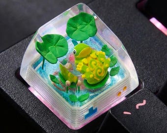 May include: A custom keyboard keycap featuring a 3D frog design. The keycap is clear with a pink and blue border, and the frog is green and yellow, sitting on a lily pad with green leaves. The keycap is on a black keyboard.