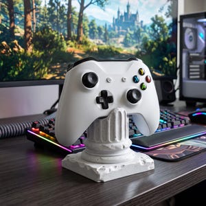 Puede incluir: Un mando de videojuegos inalámbrico blanco apoyado sobre un pequeño soporte de pedestal de piedra blanca. El mando cuenta con dos joysticks negros, una cruceta negra y cuatro botones de acción de colores etiquetados como Y, X, B y A.