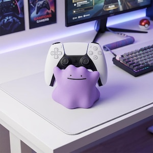 Könnte beinhalten: Ein weiß-schwarzer PlayStation 5-Controller liegt auf einem lilafarbenen Ditto-förmigen Ständer mit einem lächelnden Gesicht. Ein Computermonitor, eine Tastatur und Poster sind im Hintergrund zu sehen.