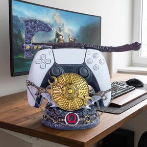 May include: A white video game controller stand featuring a decorative base with a gold circular shield, a dark axe resting across the top, and a red Omega symbol. The stand is designed to hold a controller securely in an upright position.