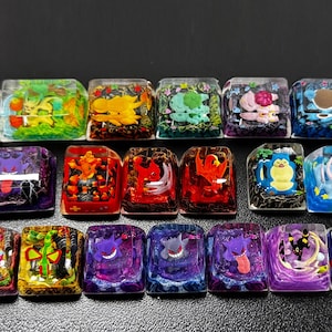 Puede incluir: Una colección de coloridas teclas de resina con varios diseños de personajes de dibujos animados. Cada tecla es de un color diferente y tiene un personaje único incrustado, creando una estética vibrante y detallada. Las teclas están dispuestas en filas.