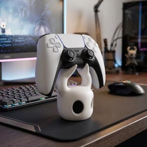 以下が含まれることがあります： 白と黒のワイヤレスゲームコントローラーが、キャラクターを模した白いスタンドに置かれています。スタンドは滑らかで丸みを帯びたベースと、コントローラーを支える2本の湾曲した突起で構成されており、コントローラーの中央には黒いタッチパッドがあります。