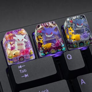 May include: Three custom artisan mechanical keyboard keycaps featuring miniature Gengar and Pikachu figures encased in clear resin. Each keycap displays a detailed, colourful diorama with purple and gold accents, set atop a black keyboard base.
