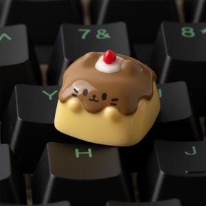 Può includere: Un copritasto personalizzato a forma di dessert, con base gialla, parte superiore marrone con una faccia carina, una macchia bianca e una ciliegia rossa. Progettato per una tastiera meccanica.