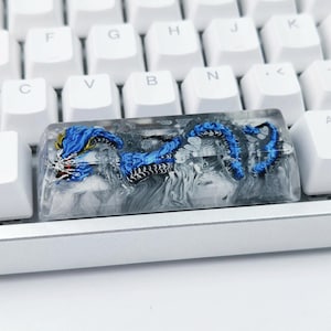 Könnte beinhalten: Eine individuelle Tastenkappe mit einem blauen Drachendesign, eingebettet in klarem Harz. Der Drache hat rote, goldene und schwarze Details. Die Tastenkappe befindet sich auf einer weißen Tastatur.