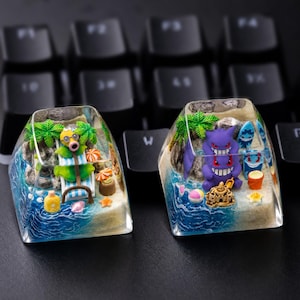 May include: Two clear resin mechanical keyboard keycaps featuring miniature beach scenes. The left keycap shows a green character on a lounge chair with palm trees. The right keycap displays a purple character, a sandcastle, and beach accessories.