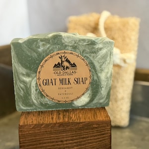 May include: A green and white marbled goat milk soap bar with a label that reads "Goat Milk Soap Bergamot & Patchouli". The soap is 4.3 oz and sits on a wooden block. A natural loofah sponge is in the background.