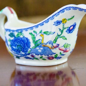 May include: A small, white ceramic gravy boat with a handle and spout. The boat is decorated with blue floral patterns, green leaves, and accents of yellow and red. The rim has a blue patterned border.