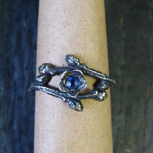May include: A silver ring with a blue gemstone set in a flower-shaped design. The ring is made to look like branches with small buds.